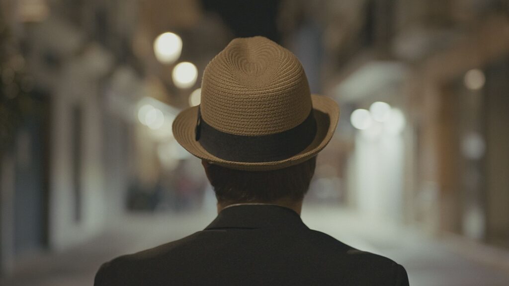 Captura del videojuego La Deuda, uno de los actores está de espaldas, con sombrero, mirando hacia la calle, se trata de una de las muchas escenas del videojuego.